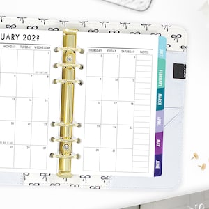 May include: A white planner with a gold coil binding and a black and white bow pattern. The planner has monthly tabs for July through June.