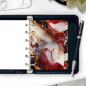 May include: A planner with a decorative cover featuring a marble design in shades of red, white, gray, and gold. The planner is open, revealing blank white pages. A dark blue pen rests beside the planner.