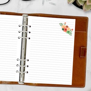 May include: A brown leather bound planner with a silver ring binder and a white lined page with a floral design in the top right corner.