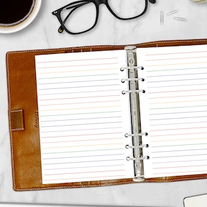 May include: A brown leather binder with lined paper featuring horizontal lines in rainbow colors. The binder is open, revealing the paper held in place by a silver, multi-ring mechanism. Black glasses and a cup of coffee are visible in the background.
