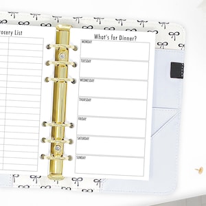 May include: A white planner with a gold binder and a black and white bow pattern on the cover. The planner has a page labeled "What's for Dinner?" with spaces to write in the meals for each day of the week.
