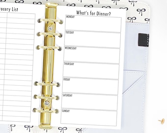 May include: A white planner with a gold binder and a black and white bow pattern on the cover. The planner has a page labeled "What's for Dinner?" with spaces to write in the meals for each day of the week.