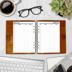 A5 bill tracker planner insert displayed open to two monthly bill tracker pages in a tan leather ring binder, styled with a black coffee cup, glasses, green succulent, and keyboard on a marble surface. Columns for Paid, Company, Due Date, and Amount. Physical paper insert only, binder not included.
