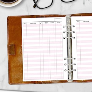 May include: A brown leather binder with a pink and white ledger insert. The ledger has columns for date, transaction, credit, debit, and balance. The binder is open, revealing the pages and metal rings.