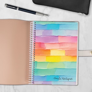 May include: Open spiral-bound notebook with a colorful abstract design. The cover features a rainbow of horizontal brushstrokes in shades of blue, purple, orange, and yellow. The name "Amala Koutapuri" is printed in a script font on the bottom right.