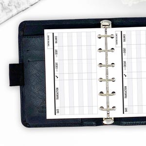 May include: A black leather binder with a silver clasp, open to reveal a white planner insert. The insert has columns labeled "Date," "Transaction," "Credit," "Debit," and "Balance." The words "Starting Balance" are also visible.