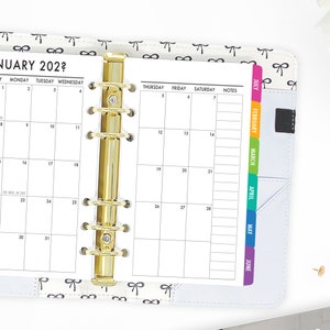 May include: A white planner with a gold coil binding and a monthly calendar for January 202?. The planner has a white cover with a black and white bow pattern. The planner has colorful tabs for each month of the year, starting with July and ending with June.