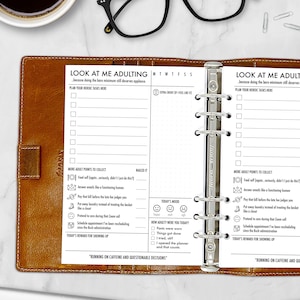 May include: A brown leather planner with a white insert featuring the text "LOOK AT ME ADULTING." The planner has a ring binder mechanism and includes sections for tasks, mood tracking, and rewards. The planner is open, revealing the interior pages.