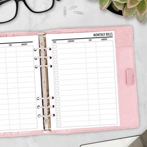 May include: A light pink six-ring binder containing white paper inserts with a printed Monthly Bills tracker. The tracker features columns for Paid, Company, Due, and Amount, with checkboxes for each entry. The binder has a metallic silver mechanism.