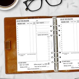 May include: A brown leather planner with the text "CEO OF THIS DISASTER" and "ONGOING CHAOS" printed on the pages. The planner is open, revealing lined pages for notes and a section for "TODAY'S PRIORITIES". Includes a section for "CEO SANITY METRICS".