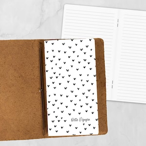 May include: A brown leather journal with a white cover featuring a pattern of black hearts. The journal is open, revealing lined pages. The name "Bette Nguyen" is printed on the cover.