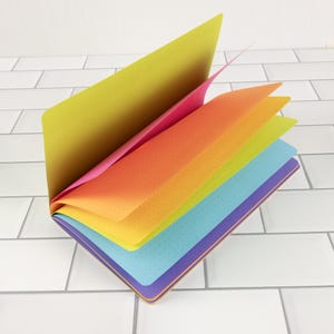 May include: A colorful notebook with pages fanned open, showcasing a gradient of colors from yellow to blue. Each page has a dotted pattern. The cover has a purple and blue border.