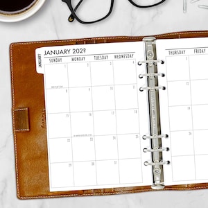 May include: A white monthly planner with a brown leather binder. The planner is open to January 2022 and shows the days of the week, with each day having a grid for writing appointments. The planner has a metal ring binder.