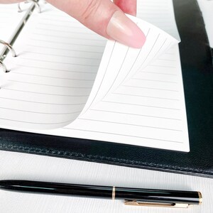 Close-up of a hand flipping through printed A5 bill tracker pages in a black ring binder, showing the clean white paper and minimalist layout. Demonstrates the quality and feel of the physical planner insert.