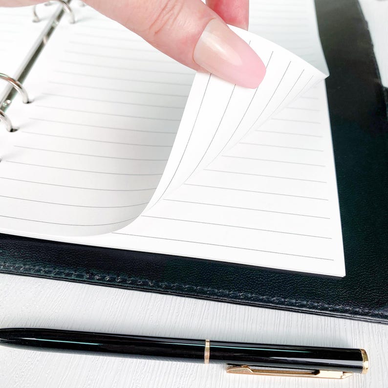 Close-up of a hand flipping through printed A5 bill tracker pages in a black ring binder, showing the clean white paper and minimalist layout. Demonstrates the quality and feel of the physical planner insert.