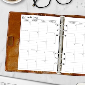 May include: Open planner with a January 2022 calendar page. The planner has a brown leather cover and a silver metal binder. The calendar is open to the month of January, with dates and days of the week clearly visible.