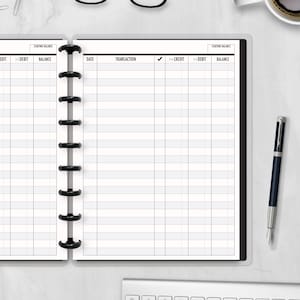 May include: A white planner with a black binding, open to a spread with lined pages for financial tracking. The left page has columns labeled "Credit", "Debit", and "Balance". A pen with a blue and silver design rests on the right side.