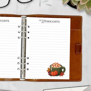 May include: A brown leather binder with a six-ring metal mechanism holding white lined paper. The top of the page features the text "cozy THOUGHTS" in black ink. A sticker of a green mug with whipped cream, a pumpkin, and leaves is attached.