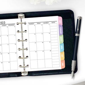 May include: A black planner with a 2023 calendar, monthly tabs, and a silver pen. The calendar pages are open to a monthly view with numbered dates and space for notes. The tabs are in various colors, including pink, green, and blue.
