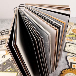 May include: An open scrapbook with black wire binding, featuring pages in various colors and textures. The pages include black, white, gray, and orange hues. Decorative elements with Halloween themes are visible around the book.
