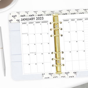 May include: A white monthly planner with a gold binder. The planner is open to January 2023 and has a bow pattern on the cover.