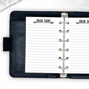 May include: Black leather planner with lined paper inserts. The paper has the words "BRAIN DUMP" printed at the top. The planner is open, revealing the metal binder rings and the lined pages.