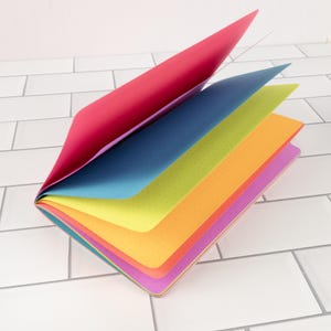 May include: A colorful notebook with pages fanned open, showcasing a spectrum of hues: red, blue, green, orange, and purple. Each page has a dotted pattern. The notebook is on a white tiled surface.