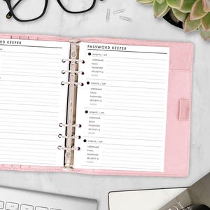 May include: A light pink six-ring binder open to a page titled PASSWORD KEEPER. The page features lined sections for website or app, username, email, password, security question, and notes. The binder has a metallic silver ring mechanism.