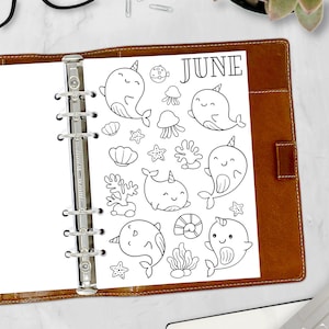 May include: A black and white planner insert for June, featuring cartoon narwhals, seashells, starfish, and coral. The word "JUNE" is printed at the top. The insert is held in a brown leather binder.