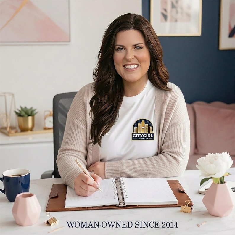 Becki, the woman owner of CityGirl Planners, smiling at a desk while writing in an open tan ring binder. She has long brown hair and wears a beige cardigan over a white tee with the CityGirl Planners logo. The desk is styled with a blue coffee mug, blush pink vases, a white peony, and gold paper clips, with a navy accent wall behind her. Caption reads WOMAN OWNED SINCE 2014. Physical paper insert only -- binder and accessories not included. Planner insert is made in the USA by CityGirl Planners