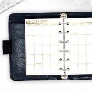 May include: A black leather-bound planner open to a January calendar page. The calendar grid is gold-colored with the text "JANUARY 20??" at the top. The planner has a silver binder mechanism and a black elastic closure.
