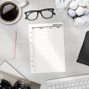 A printed planner insert for tracking monthly bills. The pages have columns for paid, company, due date, and amount. The sheet is on a white wooden surface with a cup of coffee, glasses, a pen, and a keyboard.