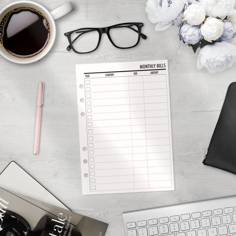 A printed planner insert for tracking monthly bills. The pages have columns for paid, company, due date, and amount. The sheet is on a white wooden surface with a cup of coffee, glasses, a pen, and a keyboard.