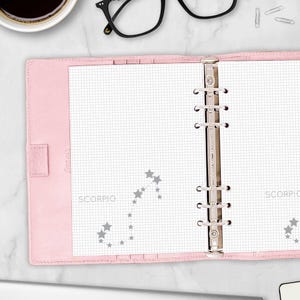 May include: A light pink personal organizer with a six-ring metal binder mechanism. The interior features white grid paper printed with a grey Scorpio constellation graphic and the text SCORPIO. The cover is a soft, matte pink material.