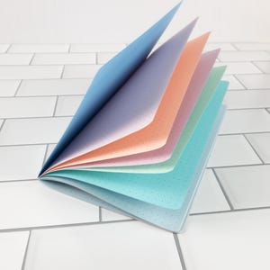 May include: A colorful notebook with pages fanned open, showcasing a spectrum of pastel hues. The pages are dotted, and the cover is a light blue. The notebook is set against a white tiled background.