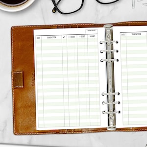 May include: A brown leather binder with a lined paper insert. The paper has columns labeled 'Date', 'Transaction', 'Credit', 'Debit', and 'Balance'. The binder is open, revealing the paper and a silver ring mechanism.