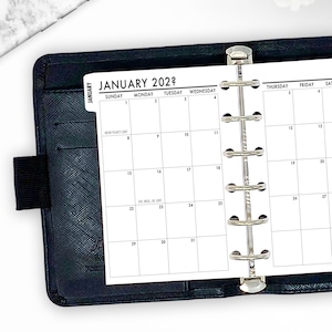 May include: A black leather-like planner with a silver metal binder. The open planner displays a January 202? calendar page with dates and holidays. The calendar is white with black text.