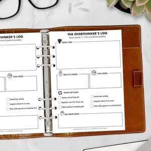 May include: A brown leather-bound planner with a metal binder. The open pages display a printed "Overthinker's Log" with sections for thought tracking, worst-case scenarios, and actual evidence. The text includes "Thought Storm" and "Final Thought (Maybe)."