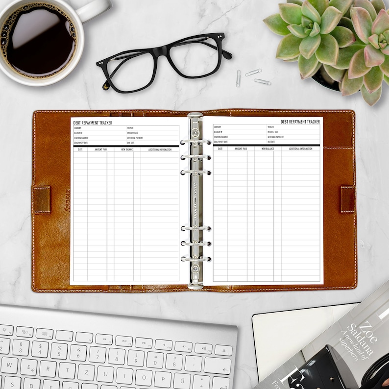 Open A5 Debt Repayment Tracker refill insert in a tan leather six-ring binder on a white marble desk. Two-page spread with DEBT REPAYMENT TRACKER header, account fields, and a payment log. Styled flatlay with a cup of black coffee, black eyeglasses, a succulent in a pink pot, a white keyboard, and a magazine. Physical paper insert only -- binder and accessories not included. Planner insert is made in the USA by CityGirl Planners