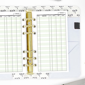 May include: A white planner with a gold binder and a light green grid pattern. The planner has columns for date, transaction, check mark, credit, debit, and balance.