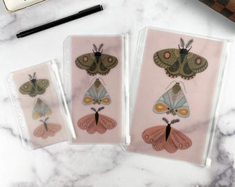 Puede incluir: Tres fundas de plástico transparente con cremallera de varios tamaños, cada una con un diseño impreso de tres polillas apiladas en tonos verde oliva, azul claro y rosa empolvado. Las fundas tienen perforaciones en el lado izquierdo.