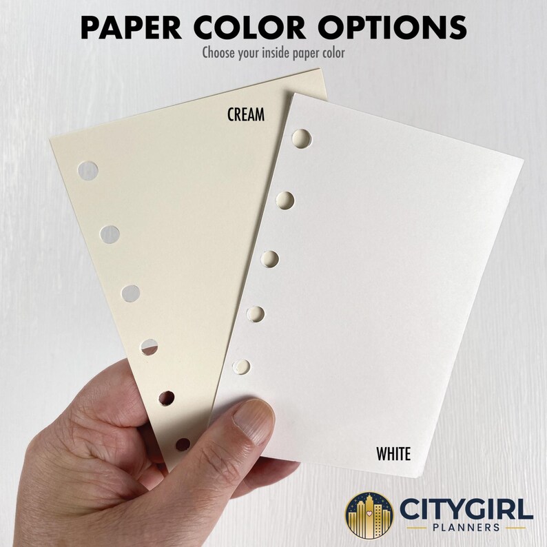 Paper color options guide showing a hand holding two punched paper samples — natural cream on the left, bright white on the right — against a light wood background. Both are 80 pound text weight, buttery smooth, and write with most pens and pencils without bleed-through. Not recommended for alcohol-based markers. Choose your inside paper color at checkout. CityGirl Planners logo displayed at bottom right.