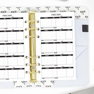 May include: A white fitness diary with a gold binder. The diary has sections for reps, meals, snacks, water, workout, and feeling. The diary is open to a page with the title "Fitness Diary".