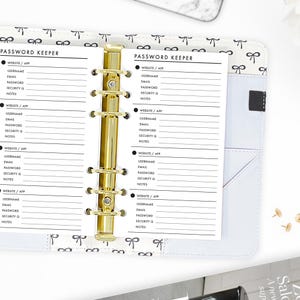 May include: An open planner with a white cover featuring a black bow pattern and a gold six-ring metal binder mechanism. The interior pages are titled PASSWORD KEEPER and contain printed fields for website, app, username, email, password, security question, and notes.