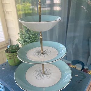 May include: A three-tiered serving stand featuring light blue and white ceramic plates with a central gray floral pattern. The tiers are connected by a central gold-toned metal rod with a circular handle at the top for carrying the stand.