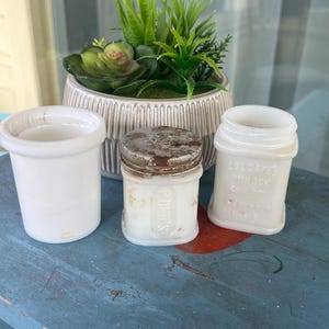 May include: Three vintage white glass jars with text, including a POND'S jar with a metal lid and a COLGATE'S MIRAGE CREAM jar. A decorative ceramic pot with green plants sits in the background on a blue painted surface.