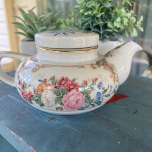 May include: A white porcelain teapot featuring a colorful floral pattern with pink, white, orange, and blue flowers. The teapot has a matching lid with a gold rim, a curved handle, and a spout, decorated with gold scrollwork accents.