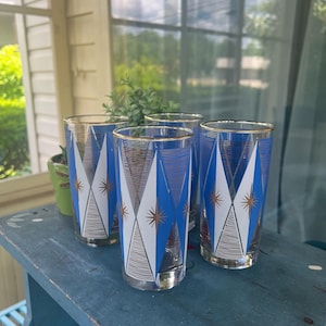 May include: A set of four clear glass tumblers featuring a repeating geometric pattern of blue and white diamonds, accented with thin gold lines and gold starburst motifs. Each glass has a gold rim and a cylindrical shape with a flat base.