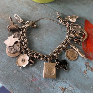 May include: A silver-toned charm bracelet featuring various metal pendants including a book, a Texas state shape with the text Texas, a Happy Birthday flower, a circular emblem, a saddle, and other assorted shapes attached to a thick chain link.