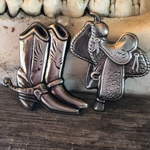 May include: Two silver-toned metal charms, one shaped like a pair of cowboy boots and the other shaped like a saddle. The charms are detailed with intricate designs.